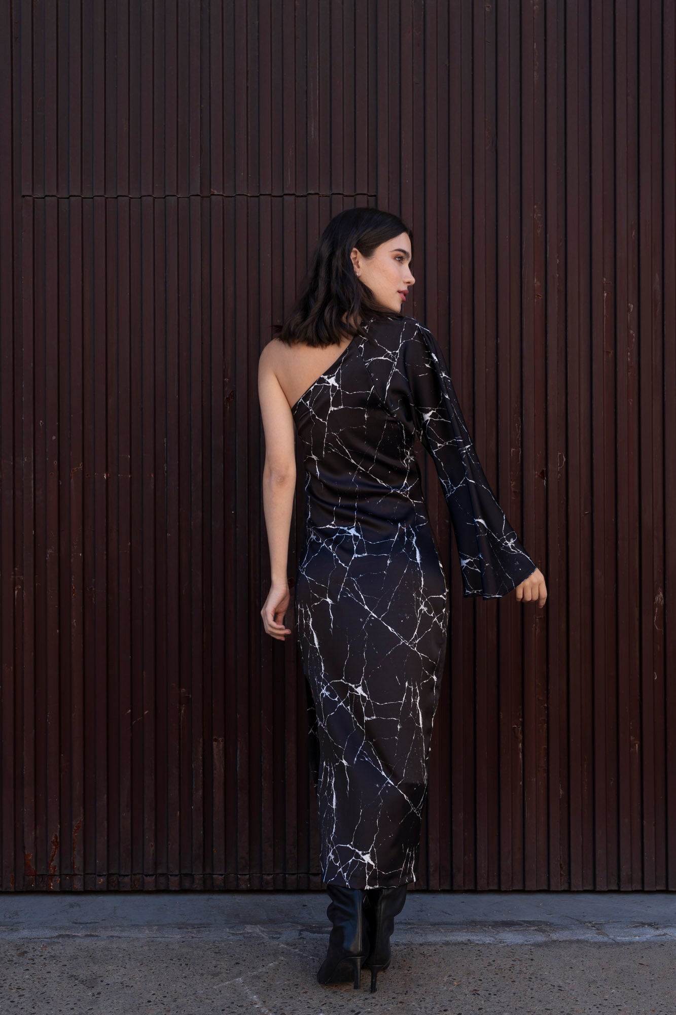 Μοντέλο που φοράει μαύρο κρεπ σατέν φόρεμα με εφέ μαρμάρου. Το φόρεμα έχει ένα μανίκι που ανοίγει στο τελείωμα και σκίσιμο στο πλάι. Η γραμμή του φορέματος είναι εφαρμοστή. Έχει συνδυαστεί με μαύρες ψηλές μπότες.