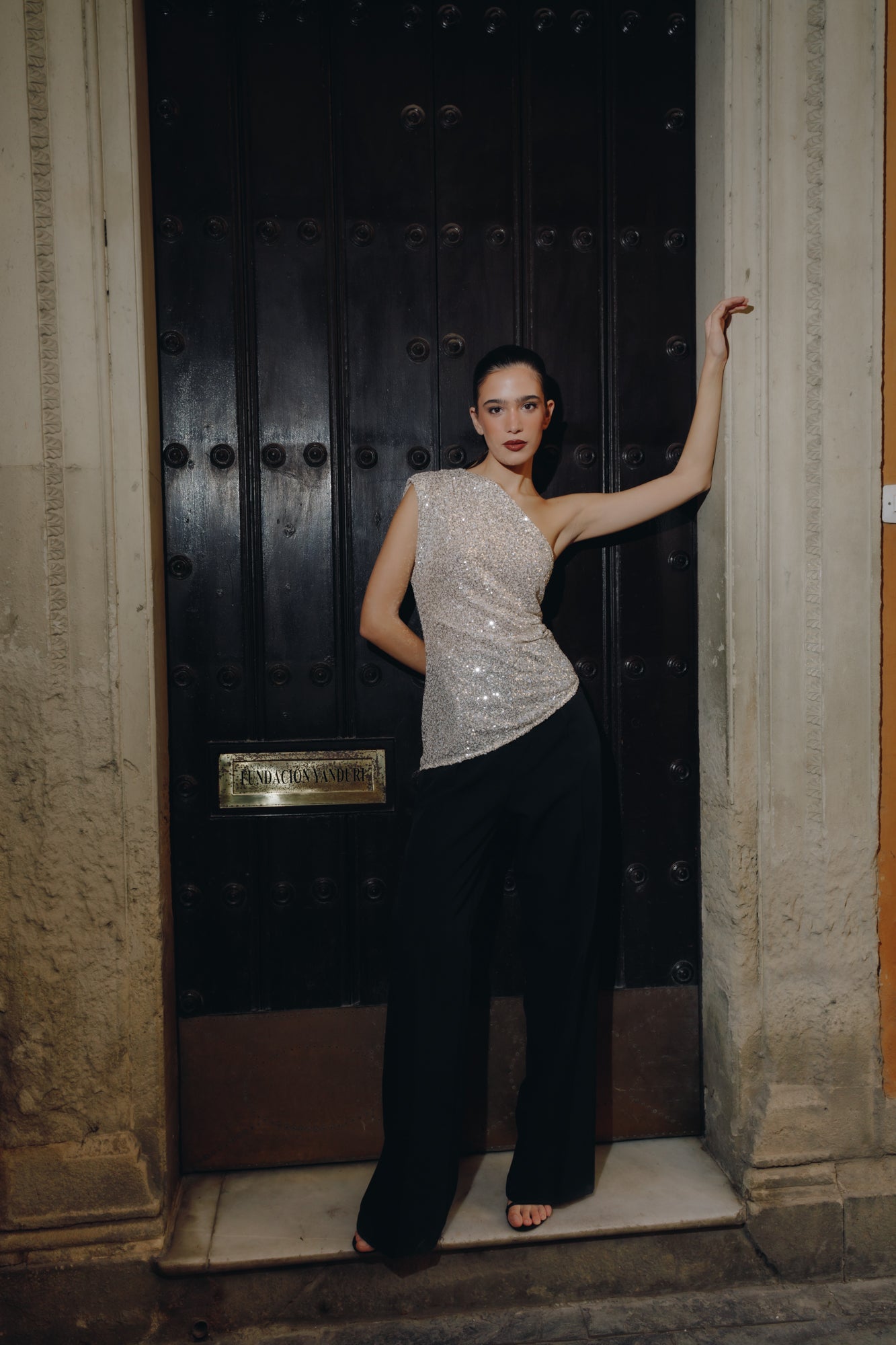 Γυναίκα που φοράει ασύμετρο τοπ από παγιέτα με έναν ώμο. Είναι ασημί και έχει σούρα στη μέση. Το έχει συνδυάσει με μαύρο παντελόνι με πιέτες.