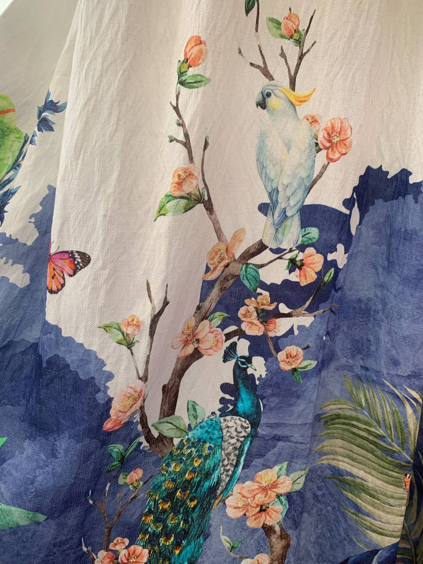 Λεπτομέρεια υφάσματος από φόρεμα που απεικονίζει έναν παπαγάλο και ένα παγώνι. Η βάση του υφάσματος είναι άσπρη και έχει πάνω μπλε αποχρώσεις και δέντρα